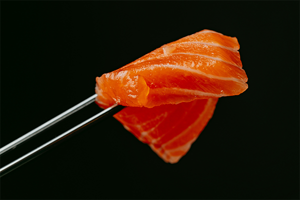 A piece of raw salmon held by metal chopsticks against a dark background.