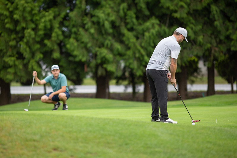 A man in a white shirt putting a golf ball on a green while another man in a green shirt watches, crouched near the hole.