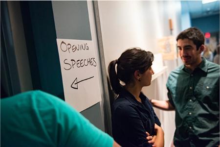 A man and a woman converse near a door with a handwritten sign saying 