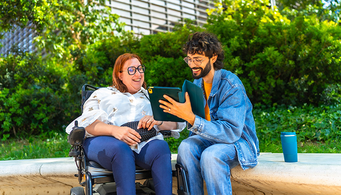 Photo of two young people sitting together, looking at an iPad and talking happily. One is using a wheelchair, while the other sits on a bench.