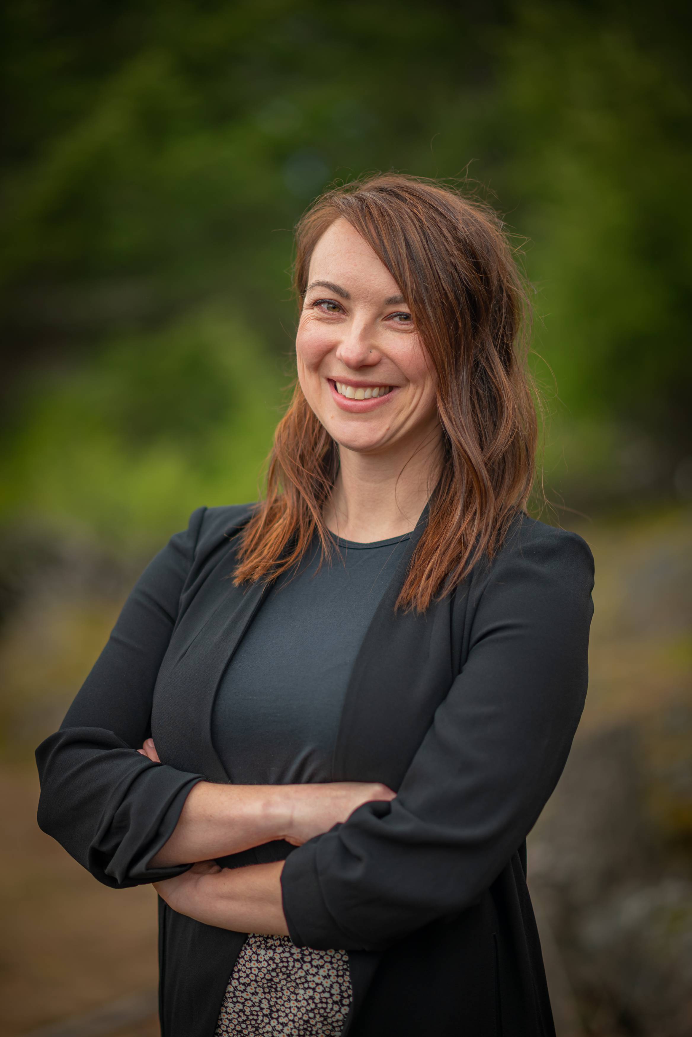 A woman standing outdoors with a blurred green, natural background. She is wearing a black blazer over a dark top.
