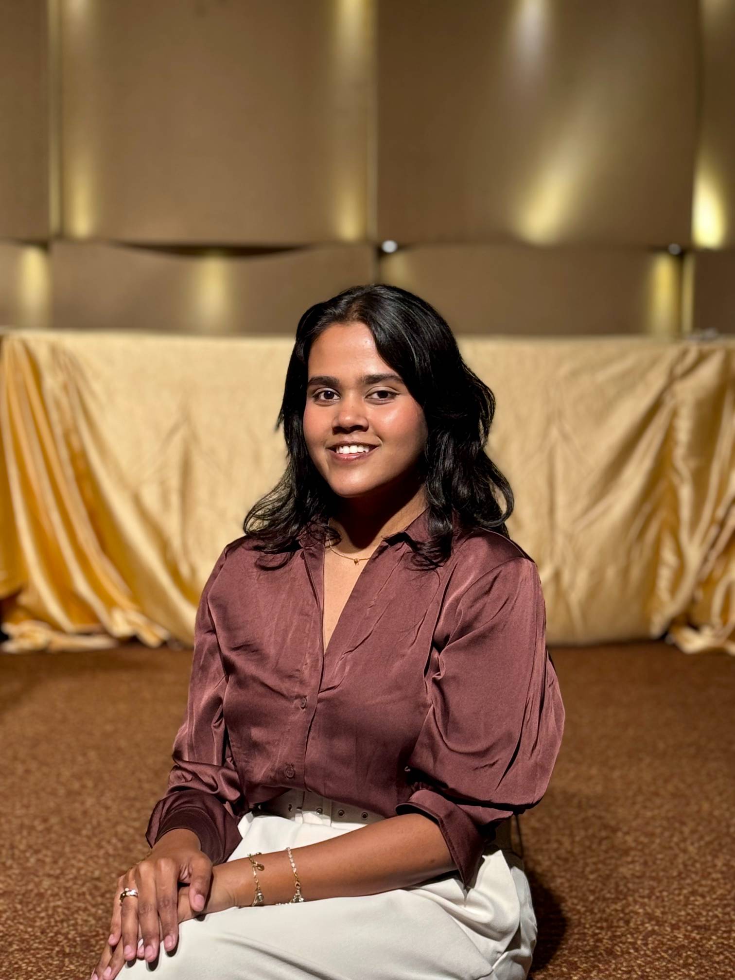 A woman seated indoors against a softly lit, warm-toned background with draped fabric and accent lighting.