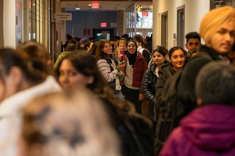 A crowd of students smiling, standing in a hallway located in building S. Focus of the photo is on a group of 2 students who are candidly smiling and facing the camera.