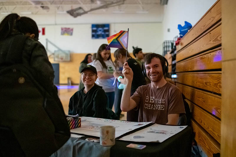 A student smiles and waves a Pride flag while welcoming fellow students to an event.