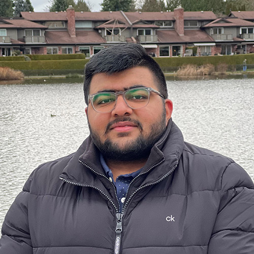 Young men dressed in a black puffy jacket looking at the camera. In the background, we can see a body of water and a row of houses.